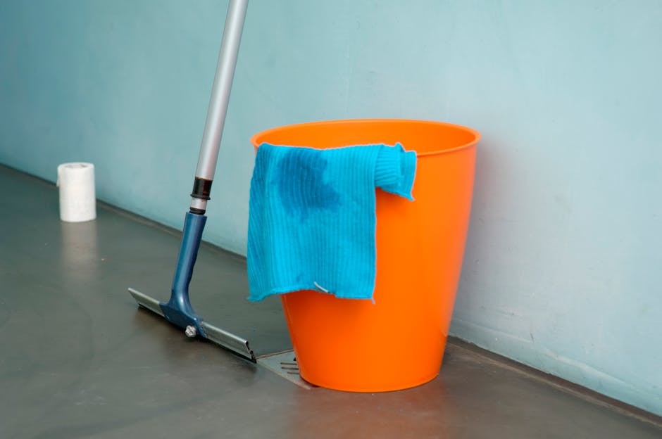 A bright orange cleaning bucket with a wet blue microfiber cloth draped over its rim, placed on a gray floor next to a drain. A silver mop with a blue handle is leaning against the bucket, and a roll of toilet paper can be seen in the background. The setting appears to be a commercial or residential space under cleaning, with clean surfaces and indoor lighting. The scene reflects surface cleaning and hygiene practices, consistent with domestic cleaning services offered by Edgware Cleaners, as highlighted in the page titled 'Avoid hidden charges from Edgware cleaning companies, EDGWARE'.