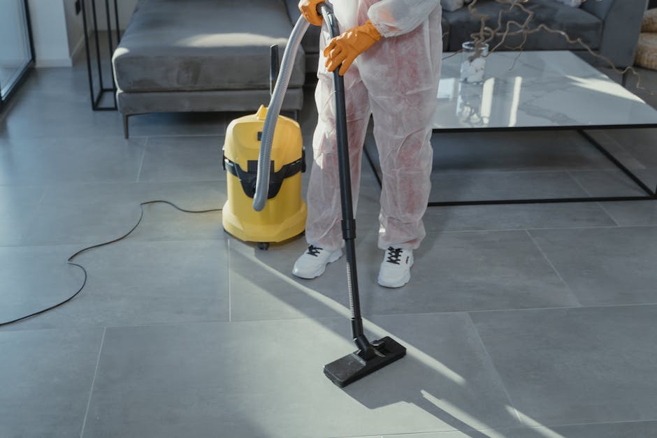 Two professional cleaners from Edgware Cleaners are standing back-to-back against a plain white wall, dressed in beige uniforms with short sleeves. The cleaner on the left has dark hair pulled back and is holding a vacuum cleaner hose in her right hand, while the cleaner on the right has light blonde hair and is holding a mop with a green handle. The scene conveys a clean, organized environment suitable for domestic or commercial cleaning, with bright, even lighting emphasizing the pristine condition of their uniforms and the uncluttered background. This image illustrates the professional cleaning services provided by Edgware Cleaners, focusing on surface cleaning and maintenance for residential or commercial spaces.