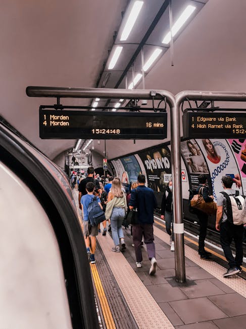 The image depicts a busy underground train station platform with passengers boarding and alighting from a modern train. The platform surface is made of smooth concrete with tactile paving strips along the edge for safety. Overhead, there are illuminated digital departure boards displaying train destinations and times, including messages related to Barnet Council waste rules for Edgware cleaning jobs. The platform has a clean and well-maintained appearance, with bright, even lighting from ceiling fixtures. Visible advertisements are attached to the train's exterior. Inside the station, Edgware Cleaners' team may utilize surface cleaning and sanitisation processes to maintain hygiene in high-traffic areas, supporting clean and safe public transportation environments. The scene emphasizes the importance of cleanliness and maintenance in busy public spaces, aligning with the service categories provided by Edgware Cleaners through their specialised cleaning and deep cleaning services.