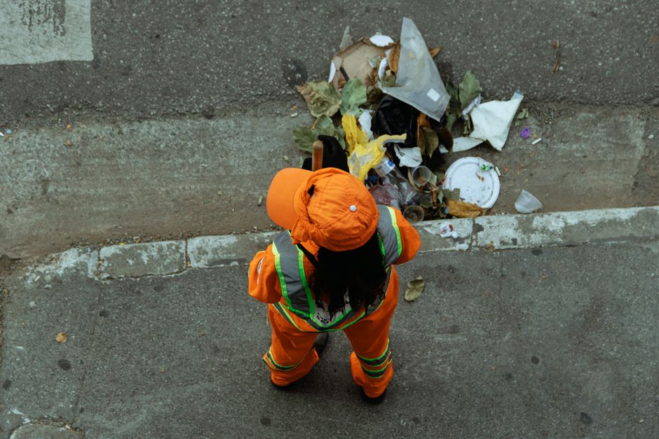 A worker dressed in bright orange high-visibility safety gear, including a jacket and a cap, is standing on a grey asphalt pavement next to a curb in an outdoor urban setting. The worker appears to be cleaning up a pile of mixed waste, such as paper, plastic, cardboard, and leaves, which is gathered against a concrete retaining wall. The surfaces around the worker are weathered, with some scattered debris on the ground. The image is well-lit with natural daylight, highlighting the contrast between the worker’s safety clothing and the waste, emphasizing the importance of sanitation and waste management in line with Barnet Council waste rules for Edgware cleaning jobs. Edgware Cleaners provides professional cleaning services focused on maintaining hygienic and safe environments through surface cleaning and waste disposal.