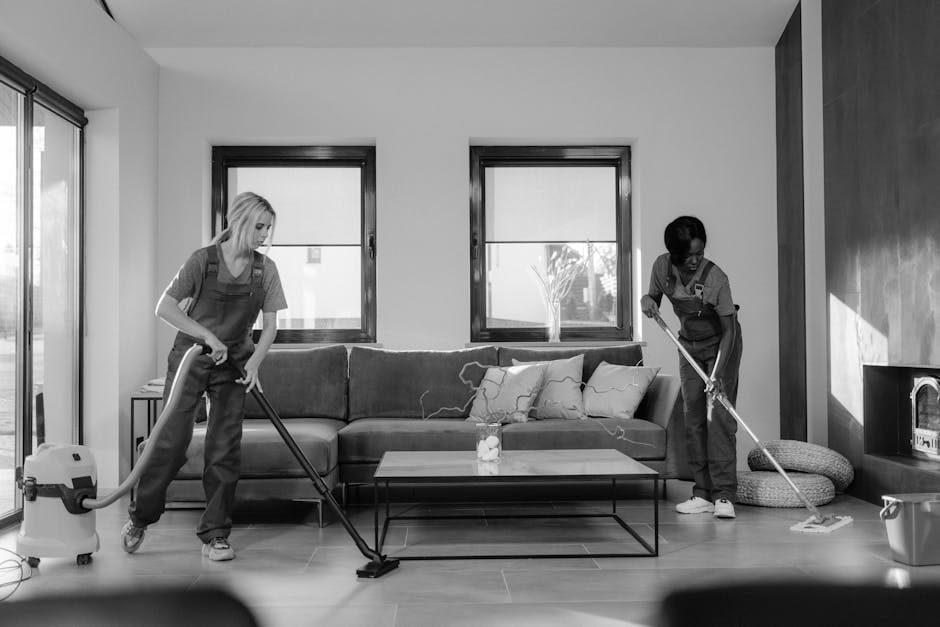Two professional cleaners from Edgware Cleaners are performing surface cleaning and deep cleaning in a modern living room. The woman on the left is using a vacuum cleaner on the wooden floor, capturing dust and debris, while the woman on the right is mopping the tiled area near a fireplace with a mop, ensuring the floor is thoroughly cleaned and sanitised. The room features a grey fabric sofa adorned with cushions, situated against a white wall with three large windows letting in natural light, highlighting the clean and well-maintained space. Visible cleaning tools include a vacuum attachment and a mop, and the room's surfaces appear dust-free and shiny, reflecting effective hygiene practices. The scene emphasizes professional domestic cleaning services provided by Edgware Cleaners, focused on surface cleaning and maintaining a hygienic living environment.