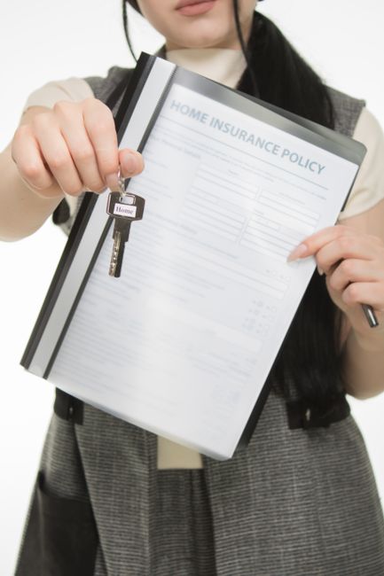 A woman with dark hair, dressed in a grey checkered blazer over a beige top, holds a folder labeled 'Home Insurance Policy' with her left hand and is handing over a key attached to a keyring labeled 'Home' with her right hand. The background is blurred, focusing on the folder and key, indicating a formal or residential setting related to property or insurance documentation.
