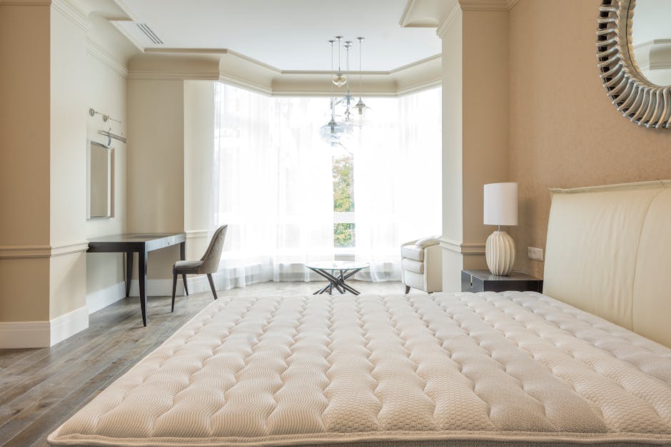 A spacious, well-lit bedroom featuring a large, beige upholstered bed with a padded headboard, positioned on a wooden floor with a clean, polished surface. To the right of the bed is a small dark beds
