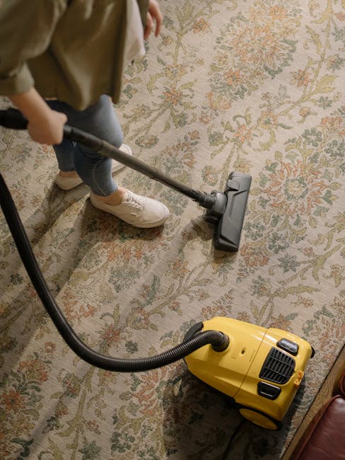 A person is demonstrated vacuuming a traditional, patterned area rug with a yellow canister vacuum cleaner. The individual, dressed in casual clothing including white sneakers and jeans, uses a hose attachment to clean the carpeted surface. The rug features a complex floral design with soft pastel colors, and is positioned on a wooden floor. The surrounding room is well-lit, highlighting the cleanliness and dust-free condition of the rug. The scene emphasizes deep cleaning and sanitisation practices, as provided by Edgware Cleaners, in preparation for same-day carpet cleaning services on Edgware Station Road and HA8.